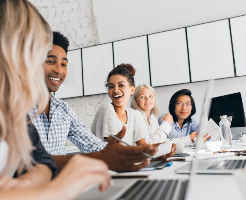 Young entrepreneurs discussing something with smile during conference. Indoor portrait of international employees sitting in office with laptops and talking about work.